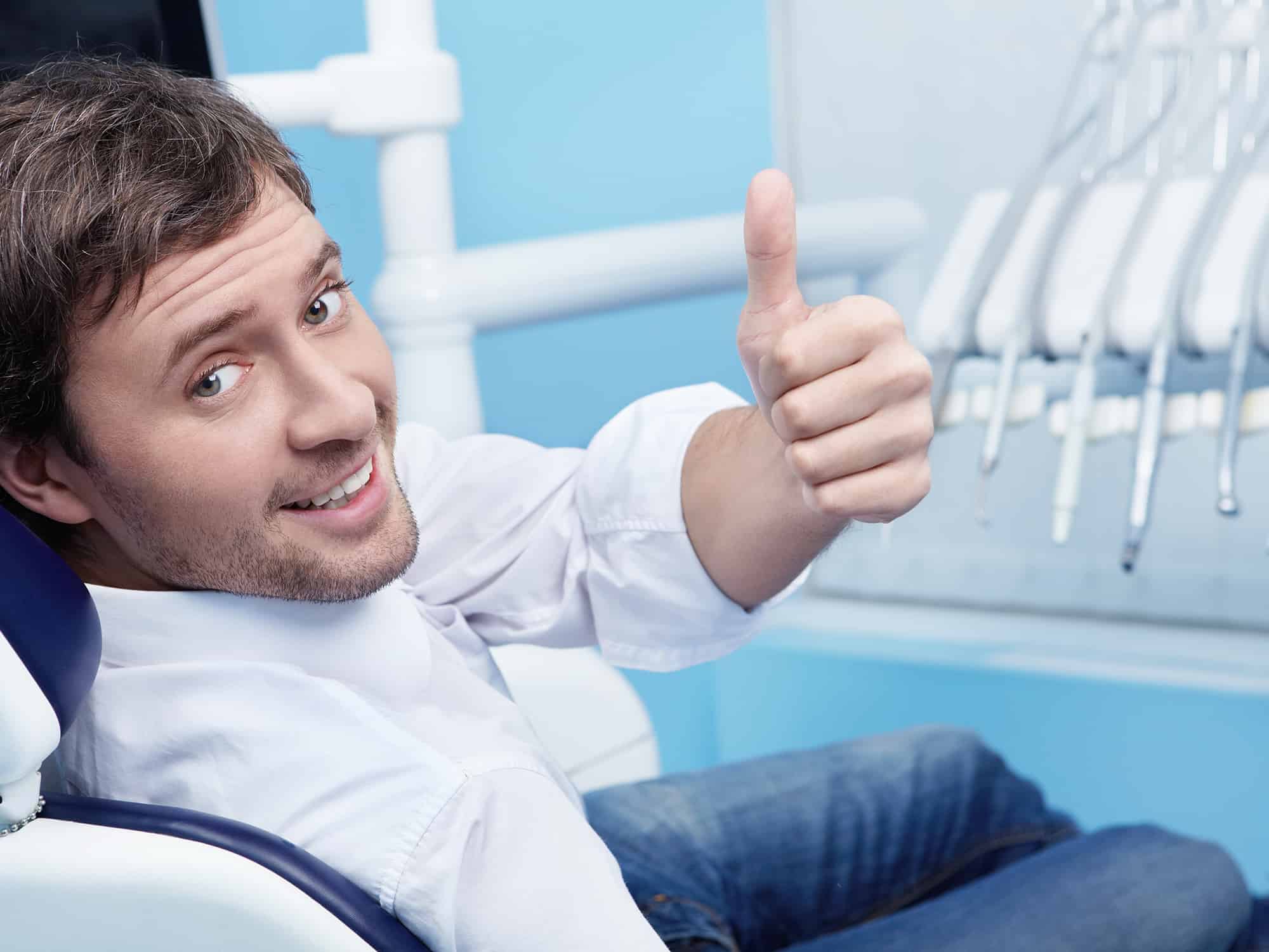  A man in a white shirt and blue jeans gives a thumbs up while sitting in a dentist's chair.